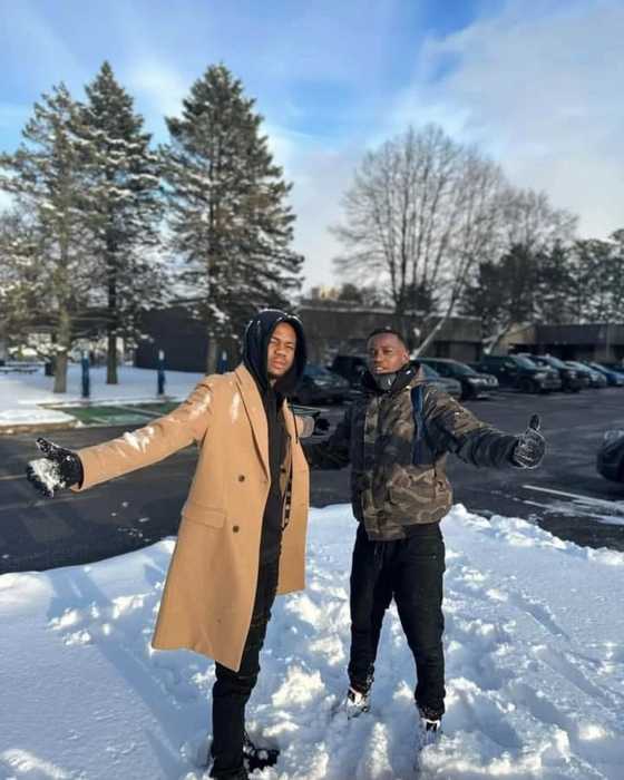 Deux jeunes hommes souriants, bras ouverts, dans la neige devant des sapins.